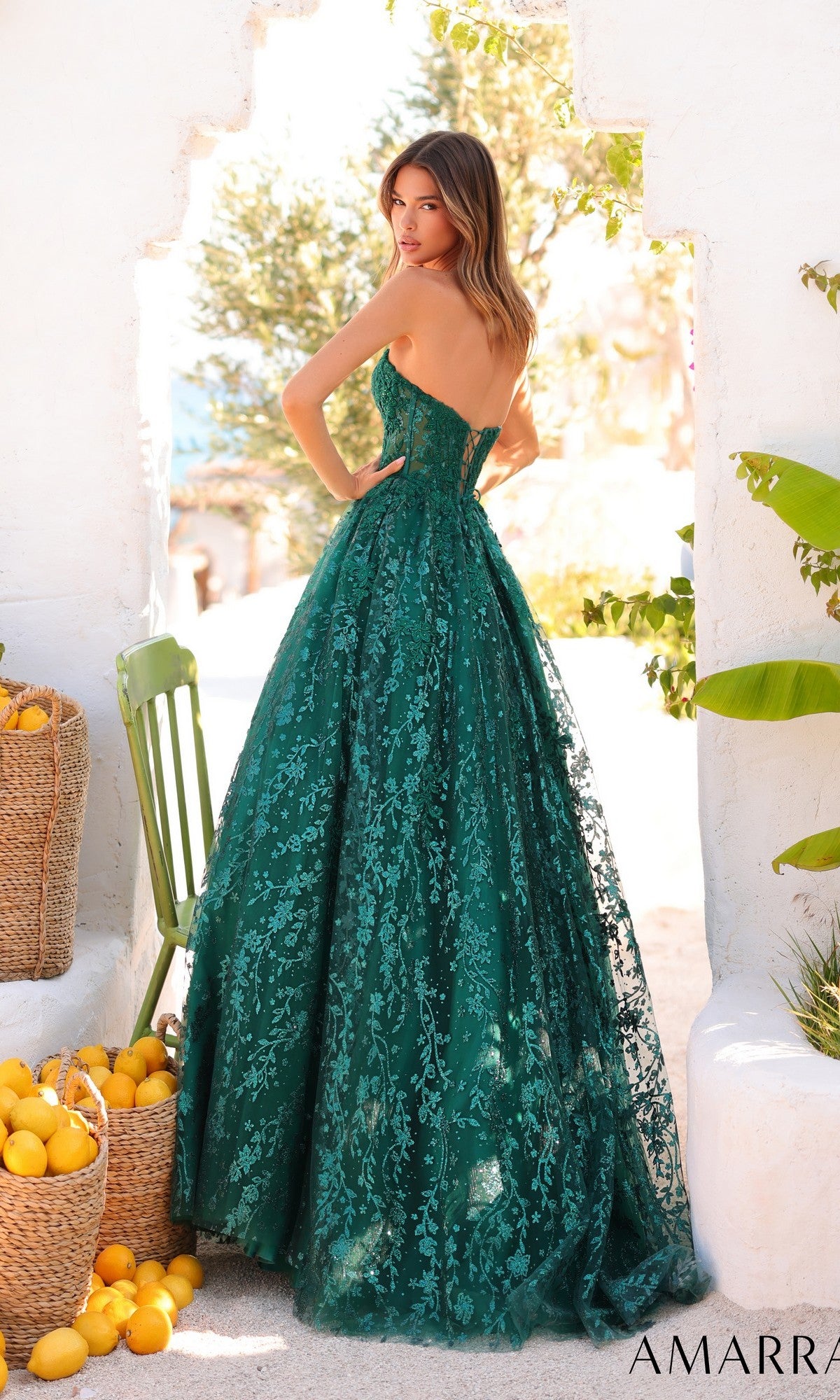 A woman in the Amarra 89427 long prom dress, featuring a strapless lace corset bodice, stands outdoors by a green chair and basket of lemons, bathed in sunlight and surrounded by lush greenery.